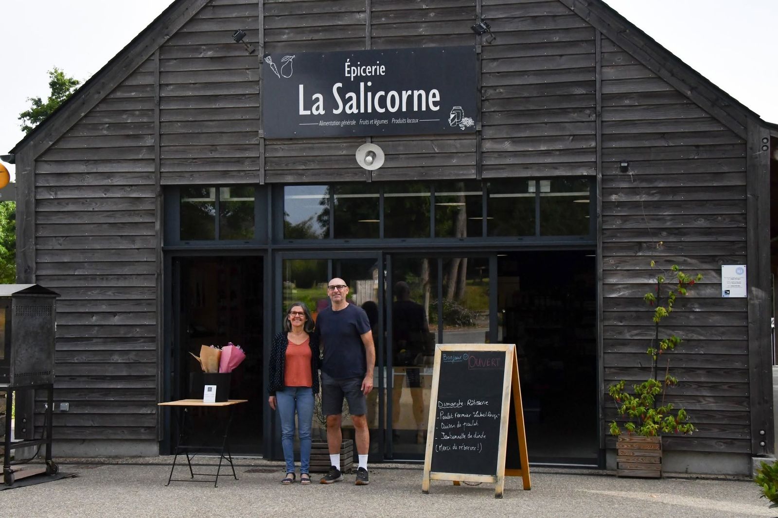 Les gérants devant l'Épicerie La Salicorne au Guerno, bâtiment en bois avec l'enseigne et l'ardoise de la rôtisserie du dimanche